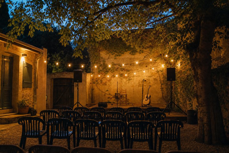 Piccolo palco allestito in un cortile al tramonto, con sedie di plastica e due casse acustiche – AI